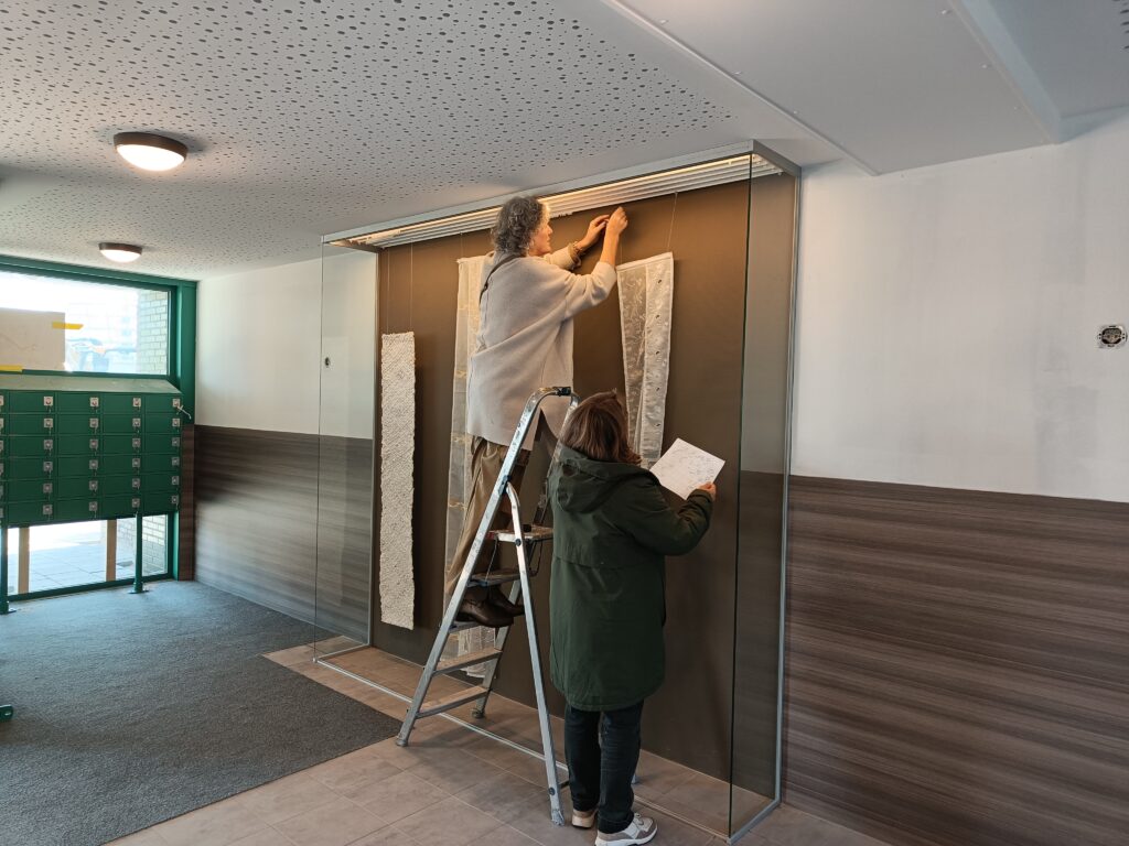 2 vrouwen die voor een glazen vitrine stroken gordijnen aan het ophangen zijn